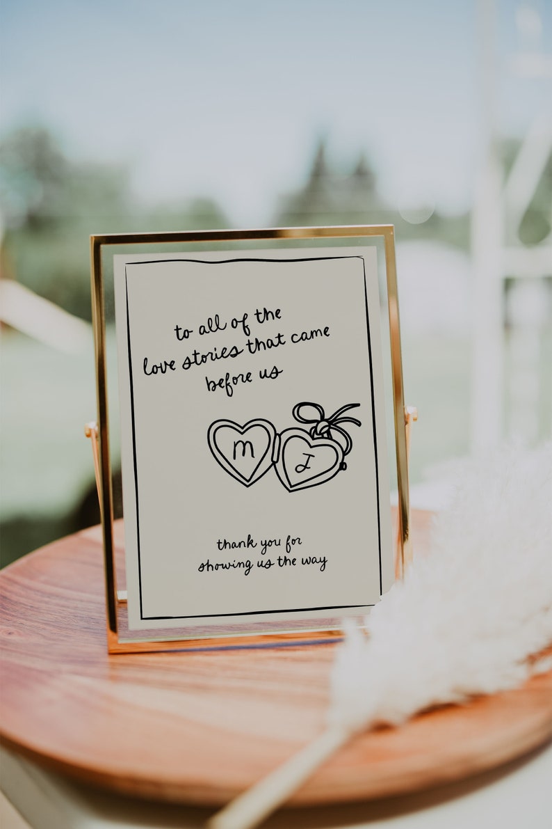 May include: A gold-framed sign with the text "to all of the love stories that came before us" and a drawing of two lockets with the letters "M" and "J" inside. The text below the lockets reads "thank you for showing us the way".