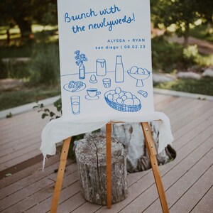 May include: A white sign with blue text and illustrations announcing a brunch with the newlyweds, Alyssa + Ryan, in San Diego on 08.02.23. The illustrations depict a breakfast table setting with food and drinks.