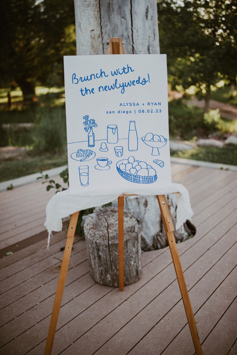 May include: A white sign with blue text and illustrations announcing a brunch with the newlyweds, Alyssa + Ryan, in San Diego on 08.02.23. The illustrations depict a breakfast table setting with food and drinks.