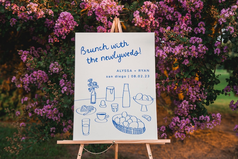 May include: A white sign with blue text and illustrations announcing a brunch with the newlyweds, Alyssa + Ryan, in San Diego on 08.02.23. The sign features a hand-drawn illustration of a breakfast spread with croissants, eggs, juice, and coffee.