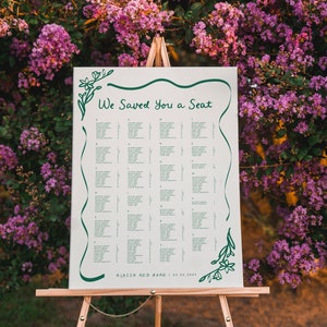 May include: A white wedding seating chart with green floral accents and the text "We Saved You a Seat". The chart is on an easel in front of a bush with purple flowers.