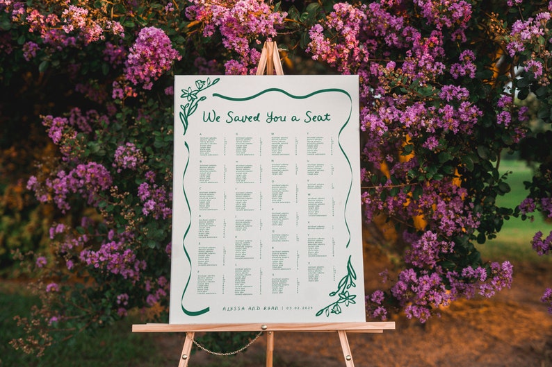 May include: A white wedding seating chart with green floral accents and the text "We Saved You a Seat". The chart is on an easel in front of a bush with purple flowers.
