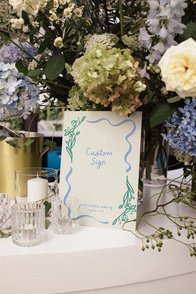 Op de afbeelding: Een wit bord met een blauwe rand en groene bloemillustraties. Het bord zegt "Custom Sign" en "Jamie Mattis Wedding August 8, 2021".