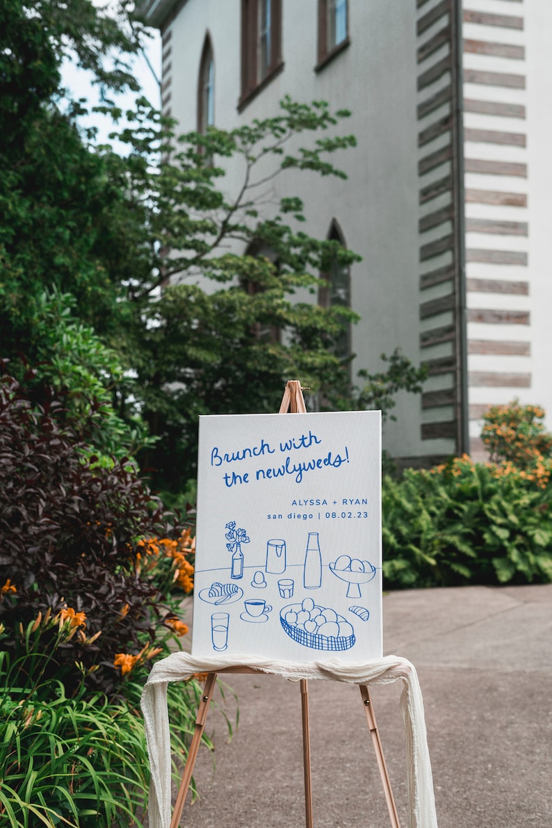 May include: A white sign with blue text that reads "Brunch with the newlyweds!"  Alyssa + Ryan, San Diego | 08.02.23.  The sign also features a hand-drawn illustration of a breakfast spread with a glass of orange juice, a coffee cup, a croissant, and a bowl of fruit.