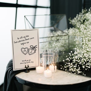 May include: A white card with black lettering that reads "to all of the love stories that came before us" with a drawing of two hearts with the initials "M" and "J" inside. The card is on a black stand and there are three clear glass candle holders with white candles on a white marble table with white flowers.