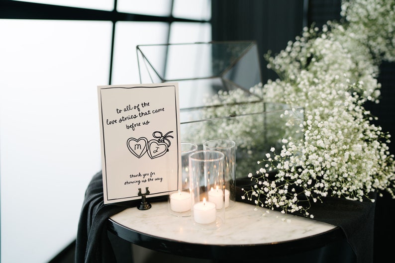 May include: A white card with black lettering that reads "to all of the love stories that came before us" with a drawing of two hearts with the initials "M" and "J" inside. The card is on a black stand and there are three clear glass candle holders with white candles on a white marble table with white flowers.
