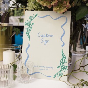 Op de afbeelding: Een wit bord met een blauwe en groene bloemenrand. Het bord zegt "Custom Sign" en "Jamie + Mattis Wedding August 9, 2021".