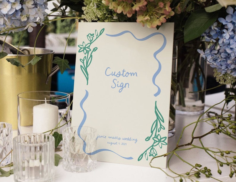 Op de afbeelding: Een wit bord met een blauwe en groene bloemenrand. Het bord zegt "Custom Sign" en "Jamie + Mattis Wedding August 9, 2021".