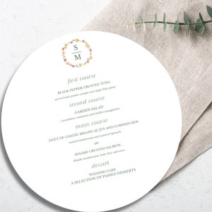 Peut inclure: Un menu rond blanc avec une couronne de fleurs et les initiales "S" et "M" au centre. Le menu propose un repas en trois services : Thon en croûte de poivre noir, Salade du jardin et au choix Duo de Chateaubriand au jus et Poulet de Cornouailles ou Saumon en croûte de sésame. Le dessert est un gâteau de mariage et une sélection de desserts servis à l'assiette.