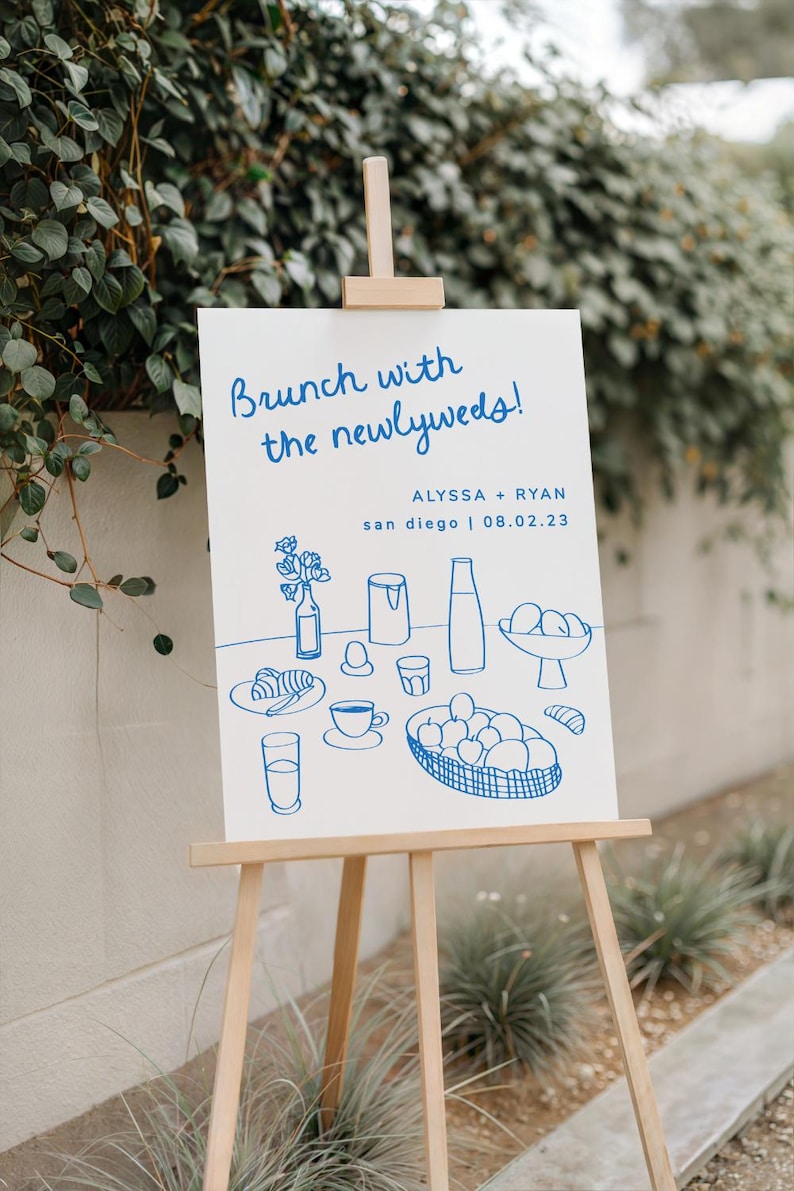 May include: A white sign with blue text and illustrations announcing a brunch with the newlyweds, Alyssa + Ryan, in San Diego on 08.02.23. The illustrations depict a breakfast scene with croissants, coffee, juice, and fruit.