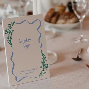 Op de afbeelding: Een wit bord met een blauwe en groene bloemenrand. Het bord zegt "Custom Sign" en "Jamie + Matty's Wedding August 4, 2023".