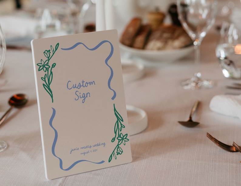 Op de afbeelding: Een wit bord met een blauwe en groene bloemenrand. Het bord zegt "Custom Sign" en "Jamie + Matty's Wedding August 4, 2023".