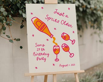 May include: A cream-colored sign on a wooden easel with the text "Just a Spritz Older" and "Sam's 27th Birthday Party". The sign features illustrations of an Aperol bottle and spritz cocktails in orange and pink.