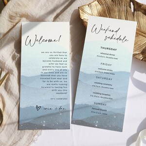 May include: Two vertical cards with a watercolor background of blue mountains and white stars. The left card says "Welcome!" and has a handwritten heart with the words "Erin & Dan". The right card says "Weekend Schedule" and lists the events for a wedding weekend, including a rehearsal dinner, welcome drinks, wedding ceremony, wedding reception, and farewell brunch.