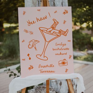 May include: Pink sign with orange text and illustrations. The text reads "Yee haw! Emily's bachelorette weekend Nashville, Tennessee". The illustrations include a cowboy hat, sunglasses, a martini glass with a woman's legs hanging out of it, a music note, and a camera.