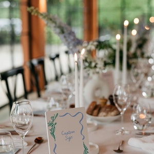 Op de afbeelding: Een wit bord met een blauwe en groene rand en bloemmotieven. Het bord heeft de tekst "Custom Sign" en "Jane & Walter Wedding August 1, 2021".