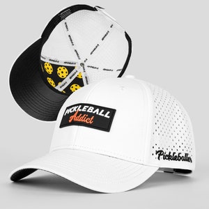 May include: White baseball cap with a black "Pickleball Addict" patch on the front and a black mesh back with "Pickleballer" printed on the side. The cap has a black brim and a white interior with a yellow and black pickleball pattern.