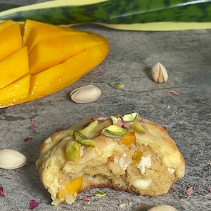 May include: A close-up of a white chocolate chip cookie with pistachios and a bite taken out of it. The cookie is on a gray surface with a few pistachio shells and pink flower petals scattered around it.