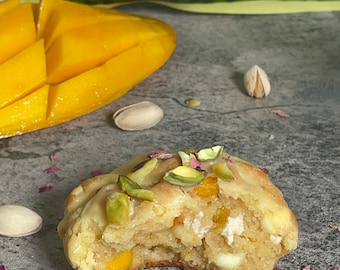 May include: A close-up of a white chocolate chip cookie with pistachios and a bite taken out of it. The cookie is on a gray surface with a few pistachio shells and pink flower petals scattered around it.