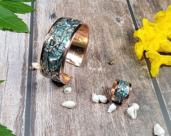 May include: A set of two copper bracelets with a turquoise blue enamel design. The bracelets are on a wooden surface with white seashells and yellow flowers.