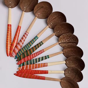May include: A collection of handmade coconut shell spoons. Each spoon features a natural coconut bowl and a wooden handle adorned with colorful, striped patterns in red, orange, green, and white. The spoons are arranged in a fan shape.