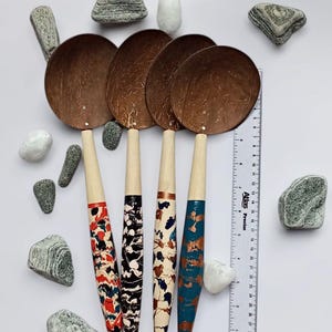 May include: Four coconut shell spoons with patterned handles in red, black, and blue. The spoons are arranged on a white surface with decorative stones and a ruler. The spoon handles are approximately 7 inches long.