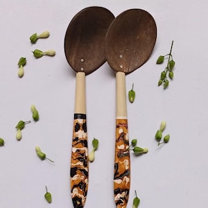 May include: Two wooden spoons with oval-shaped bowls and patterned handles in shades of brown, black, and white. The handles transition to a light wood color near the bowl. Green and white floral accents are scattered around the spoons.