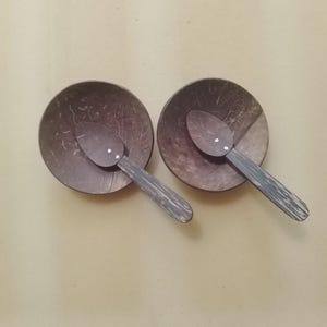 Puede incluir: Dos cuencos de coco con cucharas a juego. Los cuencos y las cucharas están hechos de un material marrón oscuro con una veta de madera natural. Las cucharas tienen un mango gris. Los cuencos son redondos y tienen un interior liso.