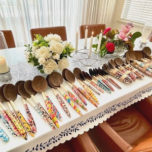 May include: A collection of wooden spoons and serving utensils with colorful floral patterned handles. The spoons have a dark brown bowl and a light-colored wooden handle. The utensils are arranged on a white tablecloth with floral arrangements and candles.