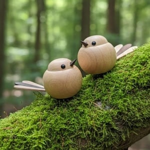 May include: Two wooden bird figurines with black beaks and eyes perch on a moss-covered branch. The birds have light brown bodies and wings made of layered wood. The background is a blurred view of a green forest.