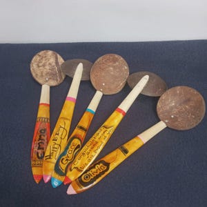 May include: Hand-painted wooden spoons with coconut shell bowls. Each spoon features a unique design and text, including names and celebratory messages. The spoons have yellow handles with colorful accents and are arranged on a dark blue surface.