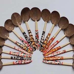May include: A collection of handcrafted coconut shell spoons arranged in a semi-circle. The spoons have light-colored wooden handles with colorful, patterned designs in red, black, and white. The spoon bowls are a natural brown color.
