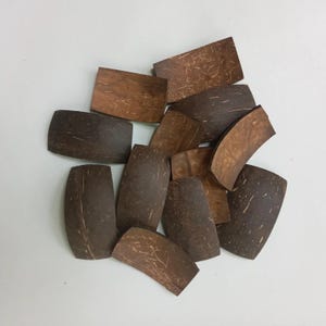 May include: A selection of dark brown coconut shell pieces in various shapes. The pieces include rectangles, ovals, and curved forms. The natural wood grain is visible on the surface of each piece, giving a textured appearance. The pieces are scattered.