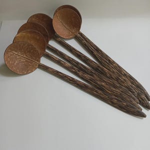 May include: A collection of seven wooden spoons with round bowls and long, slender handles. The bowls are a light brown color, and the handles have a dark brown, striped pattern. The spoons are arranged on a white surface.