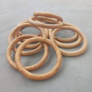 May include: A collection of light brown wooden rings, varying in size, are arranged in a loose pile. The rings have a smooth, rounded shape and a natural wood grain texture. The image is taken from a slightly elevated angle, showcasing the rings against a neutral background.