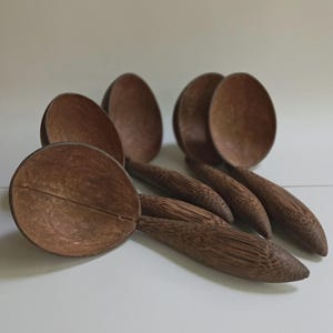 May include: A collection of six wooden spoons with round bowls and long, textured handles. The spoons are a warm brown color, and the bowls have a natural, split-like design. The spoons are arranged on a white surface.