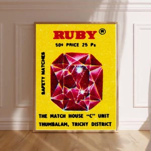 May include: A framed print featuring a vintage matchbox design. The yellow background displays the word "RUBY" in red, with "50¢ PRICE 25 Ps" below. A large, red gemstone graphic is centered. Text reads "SAFETY MATCHES" and "THE MATCH HOUSE 'C' UNIT THUMBALAM, TRICHY DISTRICT."
