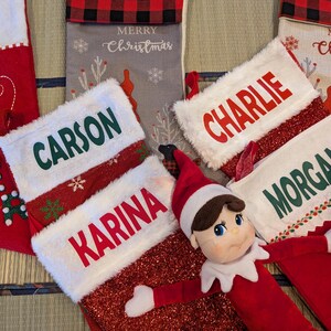 May include: Christmas stockings in red, white, and gray with personalized names: "Carson", "Karina", "Charlie", and "Morgan". The stockings have white faux fur cuffs and decorative accents. A red elf doll is also present.