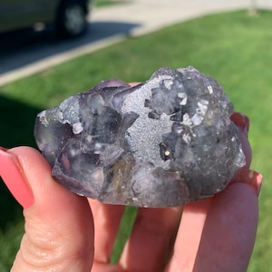 Op de afbeelding: Een ruwe, natuurlijke fluorietkristalgroep. De kristal heeft een mix van doorschijnende paarse en grijze tinten met een glinsterend, getextureerd oppervlak. De fluoriet wordt in een hand gehouden tegen een vage buitenachtergrond.