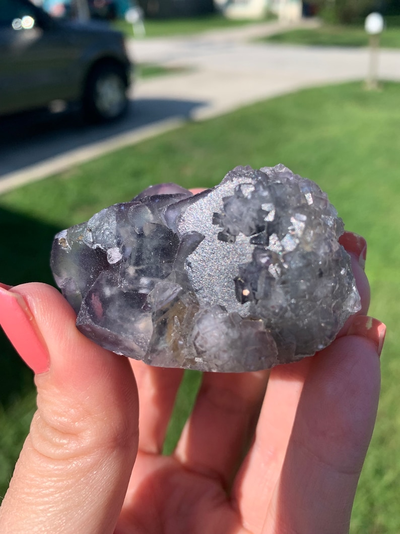 Op de afbeelding: Een ruwe, natuurlijke fluorietkristalgroep. De kristal heeft een mix van doorschijnende paarse en grijze tinten met een glinsterend, getextureerd oppervlak. De fluoriet wordt in een hand gehouden tegen een vage buitenachtergrond.