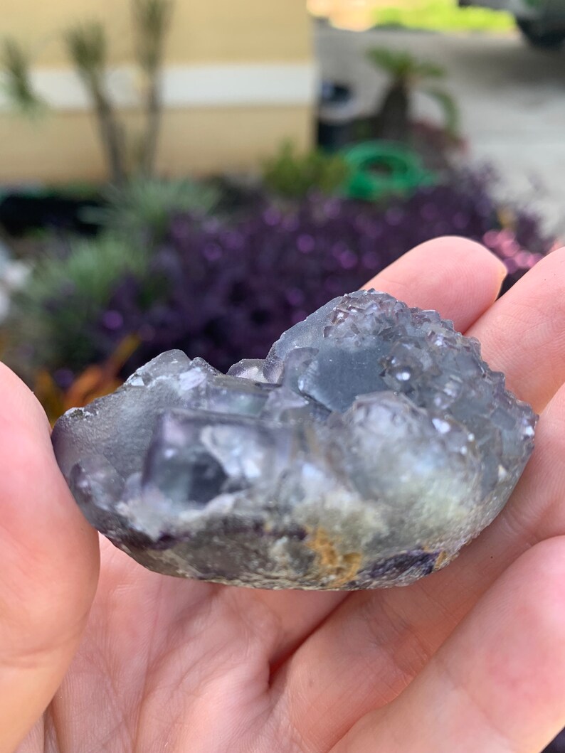 Op de afbeelding: Een kleine, onregelmatig gevormde, doorschijnende blauwgrijze fluorietkristalgroep. De kristal heeft een ruw, getextureerd oppervlak met zichtbare kristallijne structuren en een vleugje geelbruine kleuring.