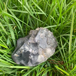 Op de afbeelding: Een ruwe, grijs-paarse kristalsteen ligt in groen gras. Het kristal heeft een facetachtige oppervlakte met doorschijnende en ondoorzichtige delen. De natuursteen is een uniek, aards item.