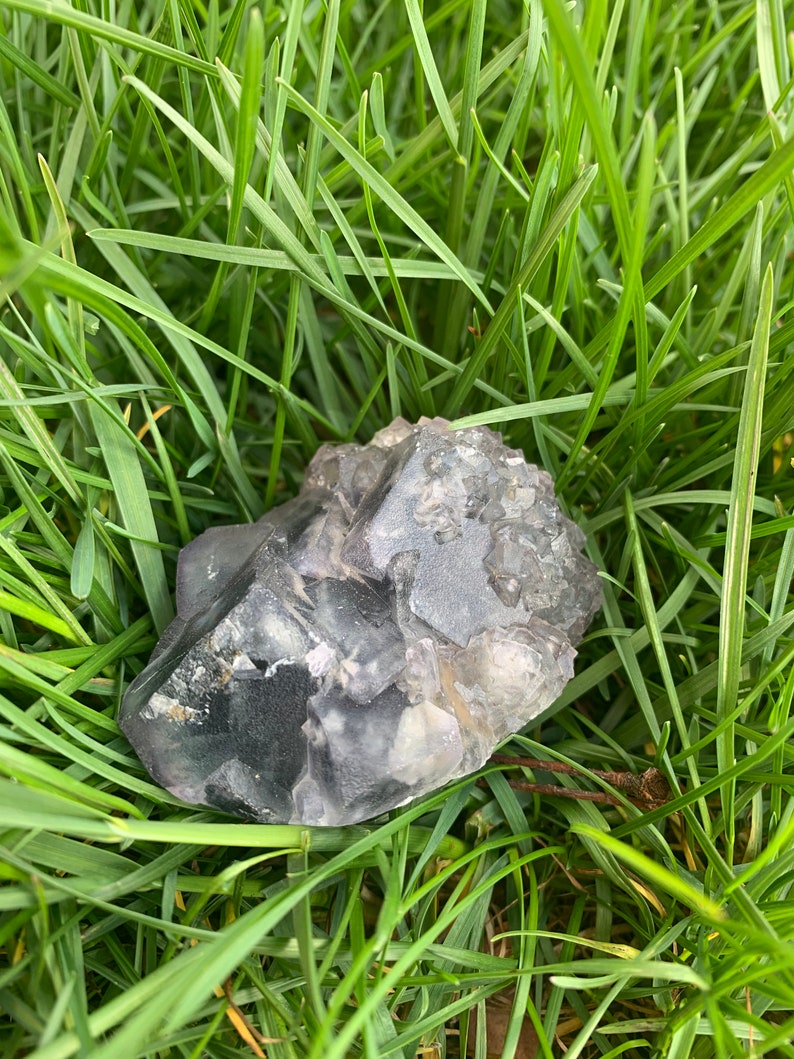 Op de afbeelding: Een ruwe, grijs-paarse kristalsteen ligt in groen gras. Het kristal heeft een facetachtige oppervlakte met doorschijnende en ondoorzichtige delen. De natuursteen is een uniek, aards item.