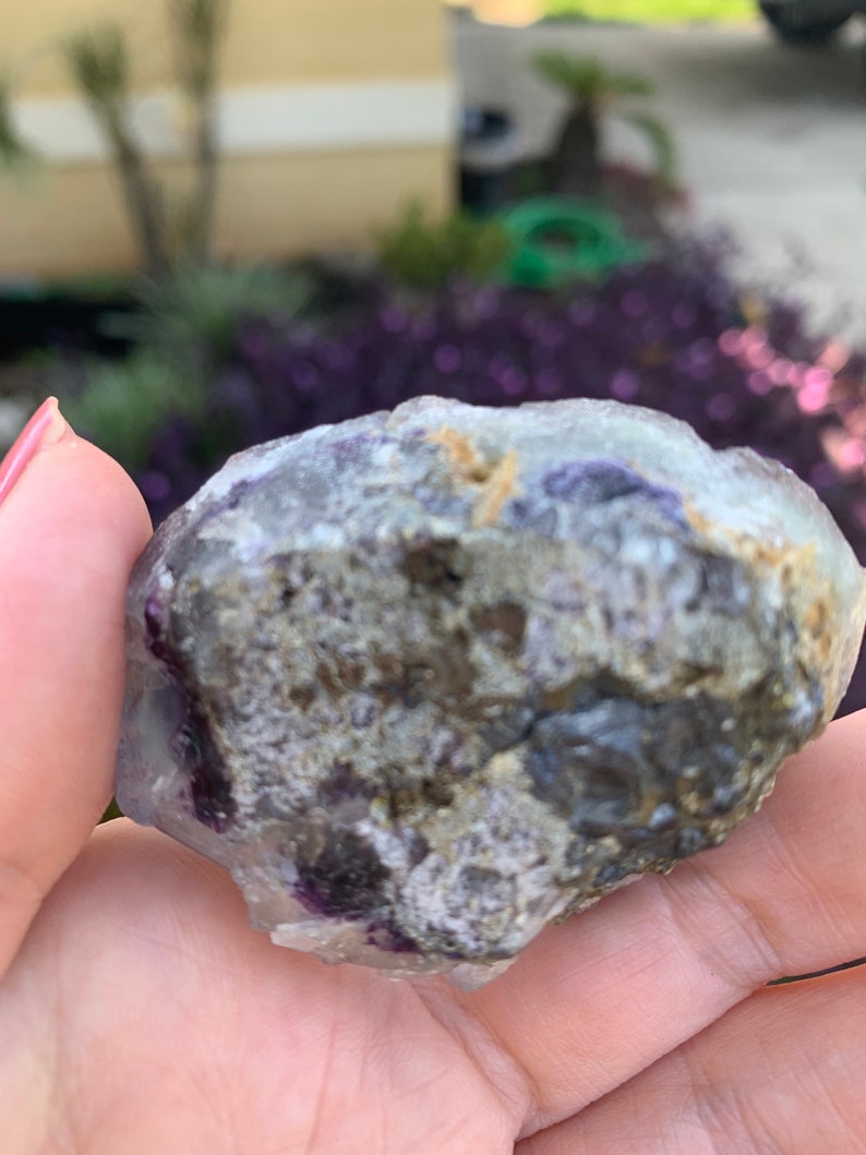 Op de afbeelding: Een ruwe, natuurlijke fluorietkristal met een mix van kleuren. De steen heeft gebieden met doorschijnend paars, grijs en bruin. Het kristal wordt in een hand gehouden, met een vage buitenachtergrond.