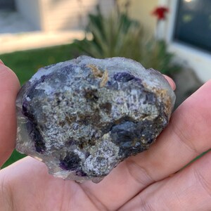 Op de afbeelding: Een ruwe, ongeslepen fluorietkristal met een mix van kleuren. De steen heeft gebieden met heldere, paarse en grijze tinten, met enkele bruine en gouden vlekken. De ruwe textuur en natuurlijke vorm zijn zichtbaar.