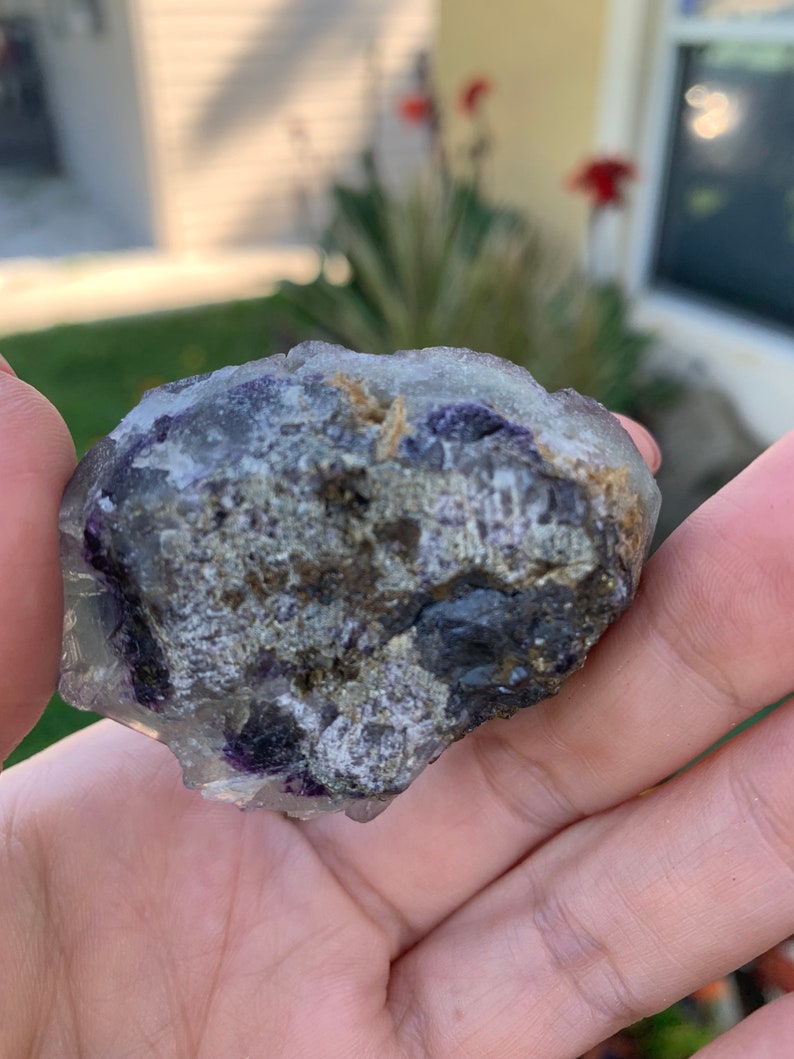 Op de afbeelding: Een ruwe, ongeslepen fluorietkristal met een mix van kleuren. De steen heeft gebieden met heldere, paarse en grijze tinten, met enkele bruine en gouden vlekken. De ruwe textuur en natuurlijke vorm zijn zichtbaar.