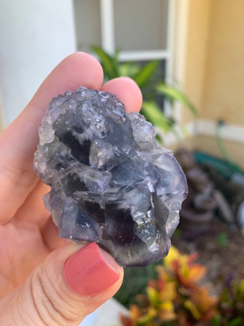 Op de afbeelding: Een ruwe, ongeslepen fluorietkristal met tinten paars, grijs en helder. De natuurlijke steen heeft een ruw, getextureerd oppervlak en wordt in een hand gehouden. De achtergrond toont groen en een gebouw.