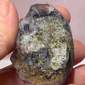 Op de afbeelding: Een ruwe, natuurlijke kristalgroep met een mix van kleuren. De steen heeft tinten paars, grijs en goud, met een ruw, getextureerd oppervlak. De kristal wordt in een hand gehouden en toont zijn natuurlijke vorm en unieke minerale samenstelling.
