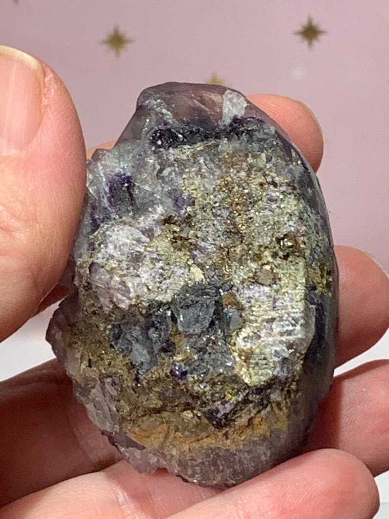 Op de afbeelding: Een ruwe, natuurlijke kristalgroep met een mix van kleuren. De steen heeft tinten paars, grijs en goud, met een ruw, getextureerd oppervlak. De kristal wordt in een hand gehouden en toont zijn natuurlijke vorm en unieke minerale samenstelling.