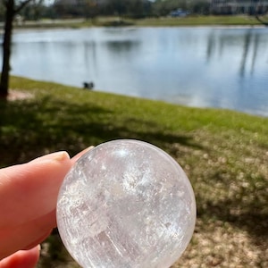 Puede incluir: Una bola de cristal transparente, esférica, sostenida en una mano. La bola tiene vetas internas y un aspecto ligeramente esmerilado. El fondo muestra un lago y follaje verde bajo un cielo soleado.